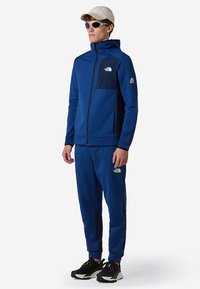 Sudadera azul y pantalones a juego hechos de tela con una textura suave. Cuenta con una cremallera frontal y detalles con logo en el pecho y la pierna.