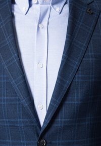 Granatowa kratkowana marynarka z jasnoniebieską koszulą w jodełkę. Posiada czarny guzik i akcent z logo na klapie.