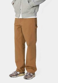 Pantalones de trabajo marrones con un corte recto, material de algodón, bolsillo lateral y costuras cosidas. Combinados con zapatillas grises y una chaqueta gris con cremallera.