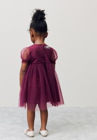 Lila tyllklänning med korta puffärmar, rynkad midja, knappstängning bak och detaljer med blommig brodyr. Bärs med ljusa skor.