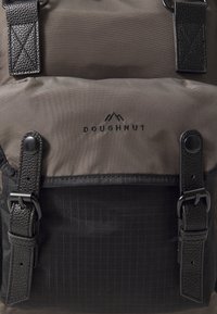 Sac à dos avec un fond noir texturé, un dessus gris, des accessoires en métal et un logo. Équipé de bretelles réglables et de fermetures à boucle.