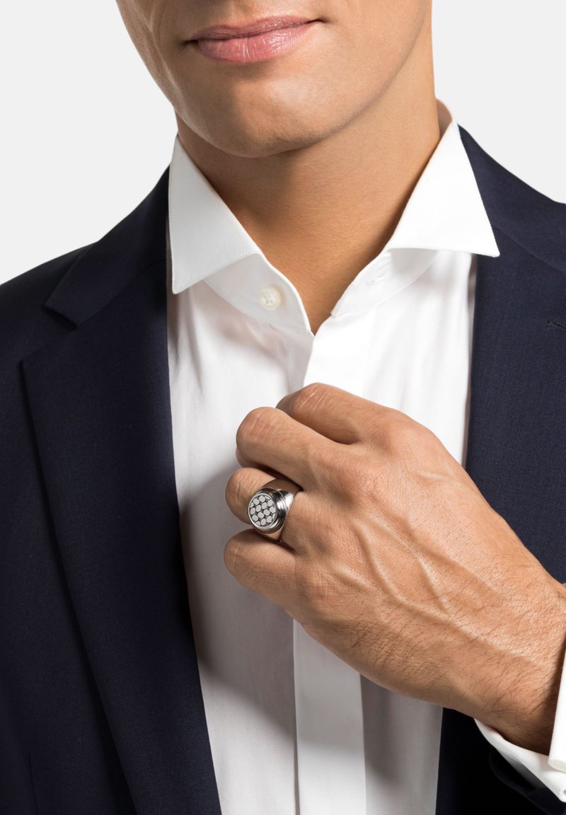 Anillo de plata con una parte superior texturizada de patrón hexagonal, llevado en una mano que ajusta una camisa blanca bajo un blazer oscuro.