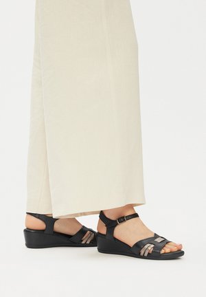 Czarne sandały na koturnie z teksturowanej skóry, z srebrnymi akcentami i paskiem wokół kostki, zestawione z jasnym, długim ubraniem.