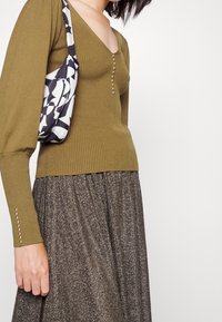 Olivgrünes geripptes Strickoberteil mit V-Ausschnitt und dekorativen Knöpfen, kombiniert mit einer schwarz-weißen mustergestalteten Tasche und einem glitzernden dunklen Rock.
