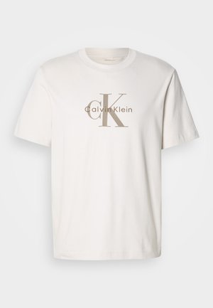 Valkoinen lyhythihainen puuvillat-paita, jossa on beige "cK Calvin Klein" -logo tulostettuna rinnan kohdalle, pyöreä kaula-aukko ja rento istuvuus.