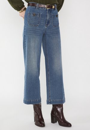 Femme portant un jean bleu taille haute à jambes larges avec poches avant et bottines en cuir marron à talons, assorti d'un haut sombre et de bracelets.