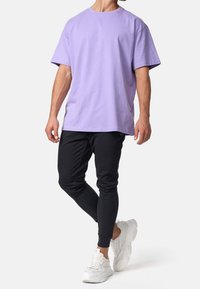 Hombre que lleva una camiseta oversized color lavanda, pantalones jogger negros y zapatillas blancas con suela gruesa, de pie con una pierna cruzada.