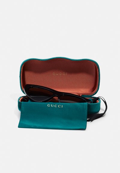 Gucci sunglasses in a black cat-eye design inside a teal hard case with a brown interior. Includes a matching soft pouch. Gold logo accents.