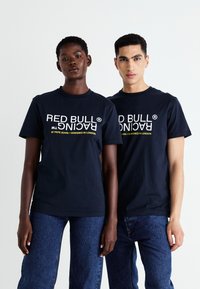 Marineblå bomulds t-shirts med "RED BULL RACING" og mærkedetaljer i hvid og gul tekst, der er parret med blå denimjeans.