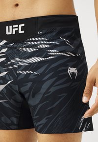 Juodos MMA šortai su dryžuotu raštu, su UFC logotipu ant juosmens ir sidabriniu dizaino elementu ant kojos.