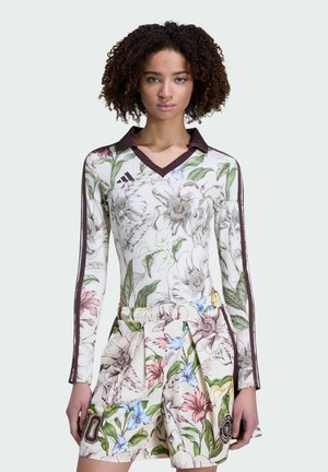 Ung kvinde iført en langærmet top med blomsterprint og matchende plisseret nederdel med mørke striber på ærmerne mod en ensfarvet baggrund.