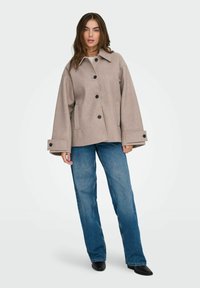 Žena u bež oversized sakou s dugmetima, plavim ravnim farmerkama i crnim špicastim cipelama, stoji protiv jednostavne pozadine.