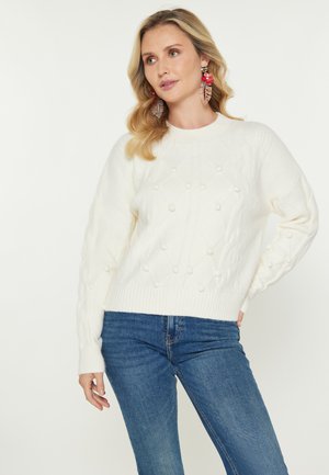 Vrouw met schouderlang blond haar, die grote bloemenoorbellen draagt, een witte textuurtrui en spijkerbroeken tegen een effen achtergrond.