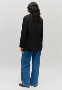 Zwarte blazer met lange mouwen, gecombineerd met losse blauwe spijkerbroek. De stof lijkt glad te zijn, met een knoopdetail op de mouw van de blazer.