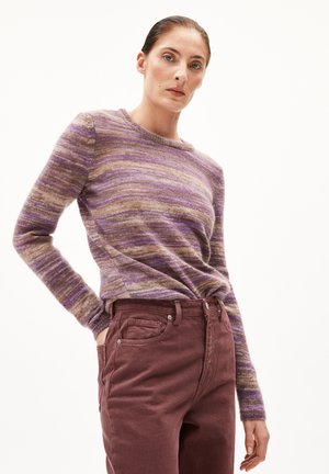 Femme portant un pull à rayures violettes et marron à manches longues, rentré dans un pantalon taille haute mauve foncé, se tenant avec une main dans la poche.