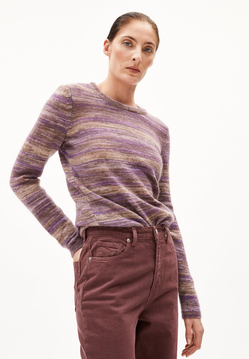 Frau in einem lila und braun gestreiften Langarmpullover, der in hochtaillierte dunkel-maulbeerpinkfarbene Hosen gesteckt ist, steht mit einer Hand in der Tasche.