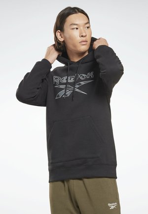 Sudadera de algodón negra con un bolsillo tipo canguro, con un logo plateado de Reebok en el pecho. Con la capucha puesta, exhibida sobre un fondo claro.