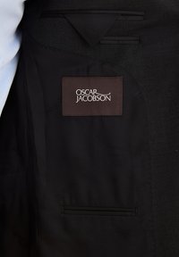 Interieur van een zwarte blazer met een pocket, voorzien van een bruine label met de tekst "OSCAR JACOBSON" in witte letters. Gladde stof en strakke lijnen.