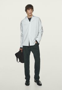 Weißes Hemd mit entspannter Passform, dunkelgrüne Jeanshose und schwarze Schuhe. Das Model hält eine braune Tasche und trägt ein schwarzes Unterhemd.