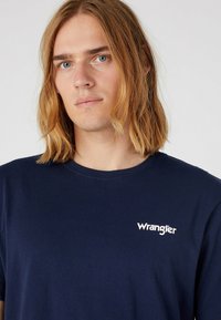 Ung person med skulderlangt blondt hår iført en mørkeblå T-shirt med hvidt "Wrangler" logo på brystet mod en ensfarvet baggrund.