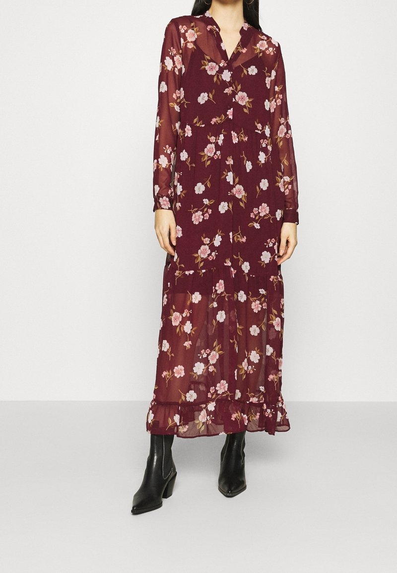 Robe maxi bordeaux à imprimé floral, manches longues, ourlet à volants et col boutonné, associée à des bottines noires.