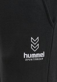 Svart sportkläder med mjukt material, prydd med vit broderad logotyp och chevronmönster. Tyget verkar slitstarkt och bekvämt.