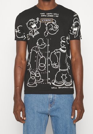 Sort t-shirt med hvide tegneserie-skitser og tekst, kombineret med blå jeans på en stående person mod en ensfarvet baggrund.