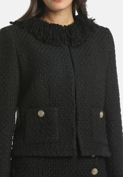 Giacca nera con texture, colletto arricciato, due tasche frontali e bottoni metallici tondi. Il tessuto presenta un motivo in rilievo e una consistenza spessa.