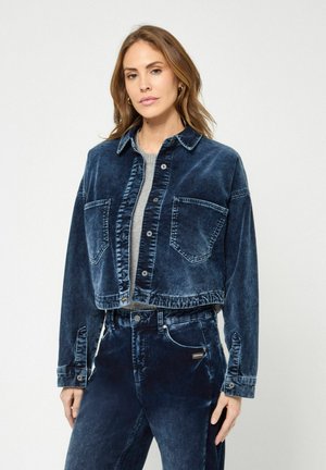 Frau mit hellbraunen Haaren trägt eine dunkelblaue, gewaschene Denimjacke und passende, hoch taillierte Jeans über einem grauen Oberteil, steht vor einem schlichten Hintergrund.