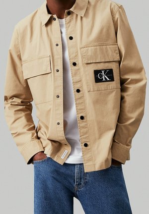 Mann trägt ein beiges Hemd mit Knöpfen und Klappentaschen und einen schwarzen Calvin Klein Jeans Logo-Patch, weißes T-Shirt und blaue Jeans, Hände in den Taschen.