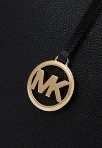 Schwarze, strukturierte Lederhandtasche mit einem auffälligen, runden, goldfarbenen Logo-Anhänger mit den Initialen "MK". Glatte Akzente kontrastieren mit der strukturierten Oberfläche.