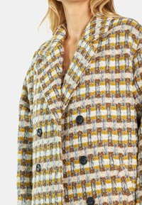 Dwurzędowy płaszcz wykonany z teksturowanej tkaniny o splocie w żółtym, brązowym i białym kolorze, z czarnymi guzikami i szerokim kołnierzem.