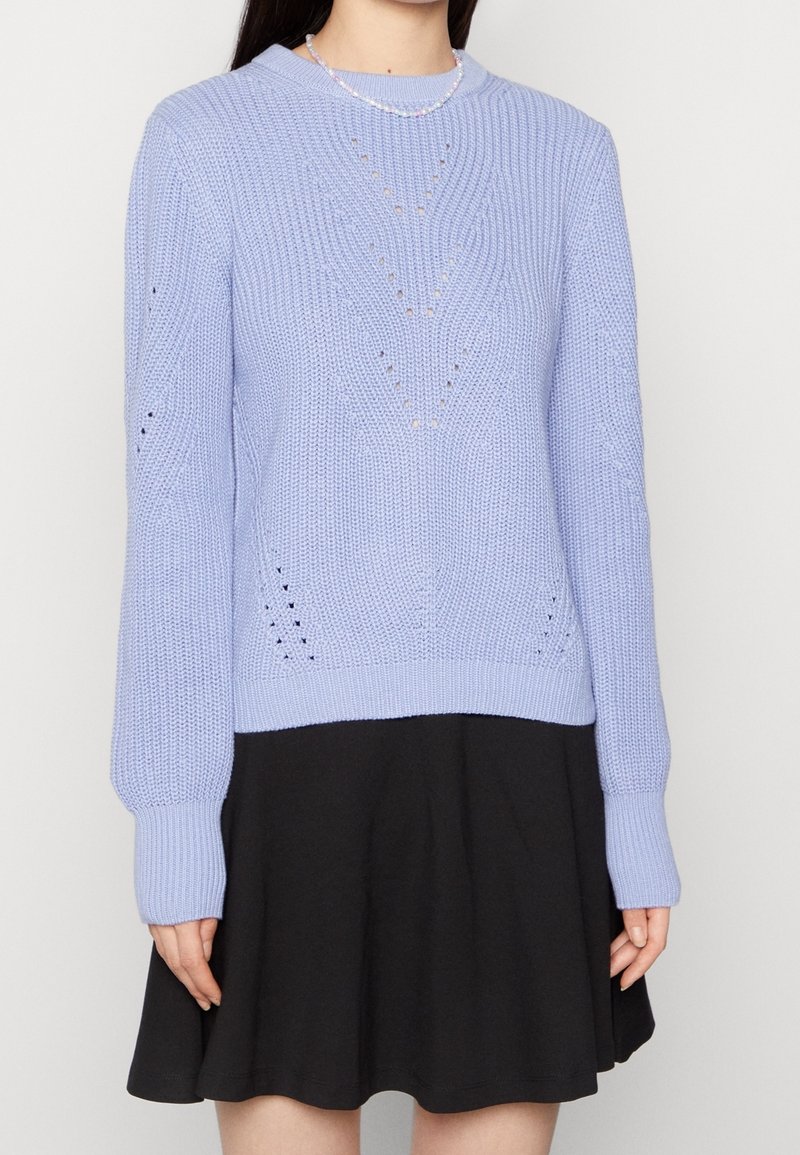 Suéter de punto azul claro con un patrón texturizado y detalles de calado. Presenta mangas largas y un cuello redondo. Combinado con una falda negra.