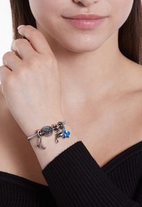 Zilveren armband met bedels, waaronder een blauwe steen, een dolfijn en bloemaccenten, gedragen om een pols. Textuur van een zwarte top zichtbaar.