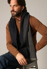 Écharpe marine avec un motif texturé aux accents beiges, finie par des bords frangés. Portée par-dessus un pull zippé marron.