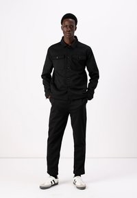 Camisa preta de botão com dois bolsos no peito, combinada com calças pretas e ténis brancos com riscas pretas, de pé contra um fundo simples.