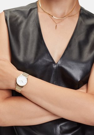 Montre-bracelet en or avec un cadran blanc et un bracelet en maille texturée, portée sur un bras nu. En arrière-plan, un vêtement en cuir noir brillant.