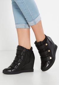 Svarta kilskor med justerbara kardborreband, glansig yta, rund tå, sidodragkedja och texturerad sula. Bärs med uppvikt jeans.