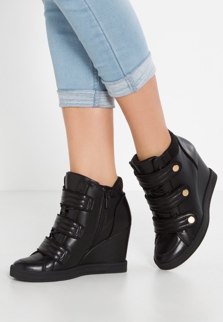 Svarta kilskor med justerbara kardborreband, glansig yta, rund tå, sidodragkedja och texturerad sula. Bärs med uppvikt jeans.