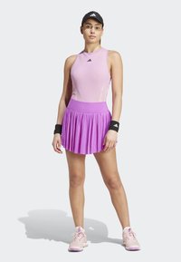 Roze sportieve tanktop en plooienrokset. De rok heeft een brede paarse tailleband. Zwarte polsbanden en roze sportschoenen maken de look compleet.