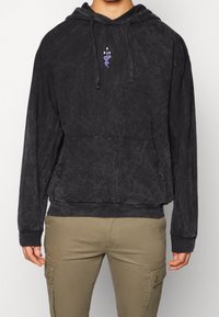 Sort hættetrøje i blødt, tekstureret stof med et lille lilla grafisk design på brystet, med en kængurulomme og ribstrikkede ærmeafslutninger.