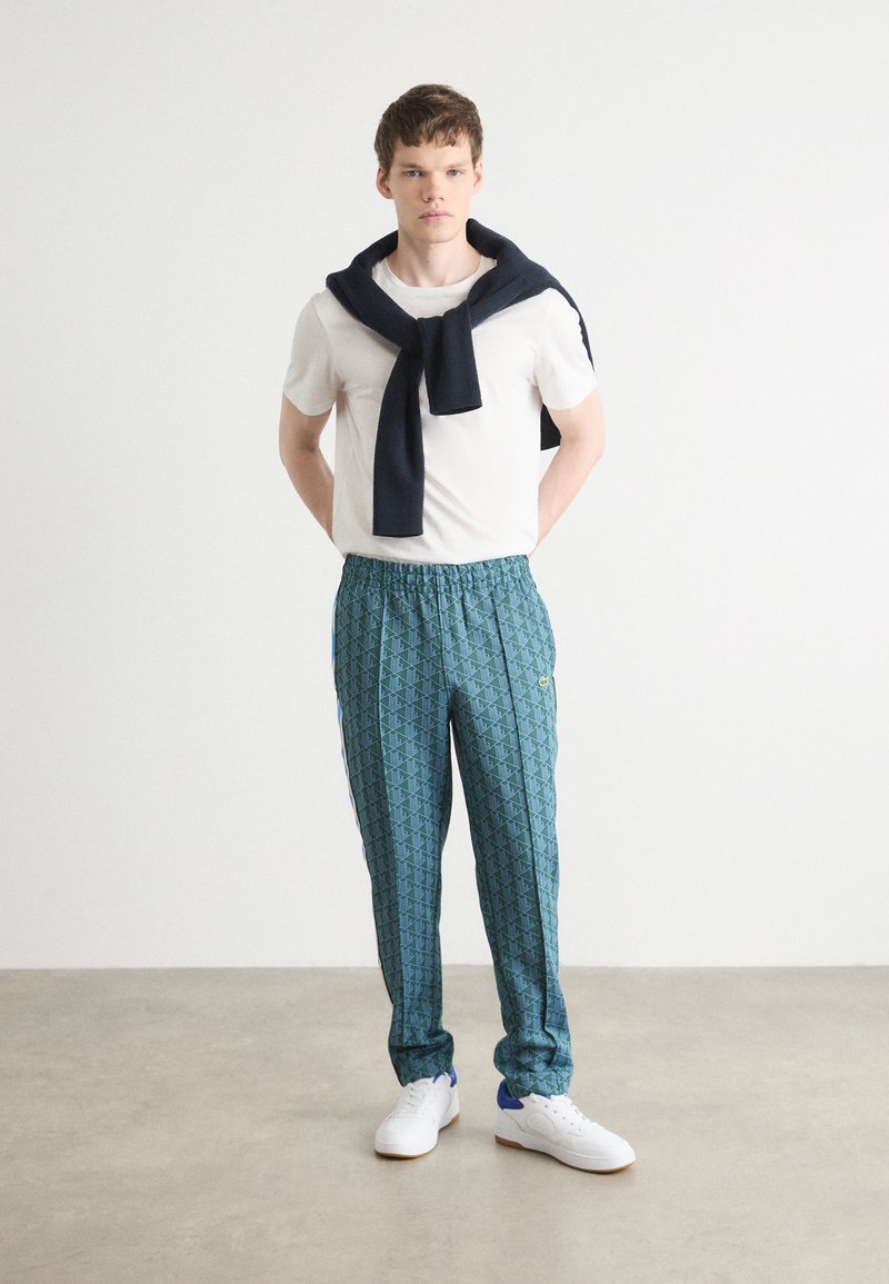 Jogging vert à motifs avec un design géométrique, associé à un t-shirt blanc et un pull marine foncé noué aux épaules. Des baskets blanches complètent le look.