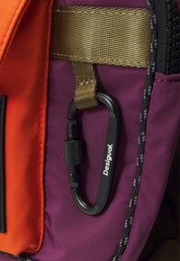 Mousqueton noir avec le texte "Desigual" attaché à un sac en tissu violet et orange avec une sangle marron et des bords de fermeture éclair noirs.