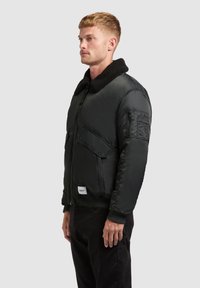 Jachetă bomber pentru bărbați, de culoare neagră, cu guler pufos, buzunare cu fermoar, manșete ribbed și un finisaj de țesătură netedă și lucioasă.