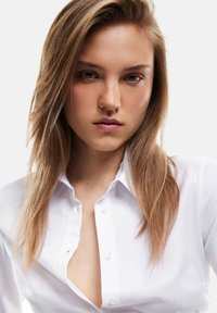 Una camicia bianca con colletto, caratterizzata da un collo leggermente aperto e una trama di tessuto morbido. I capelli biondi lisci incorniciano parzialmente il viso.