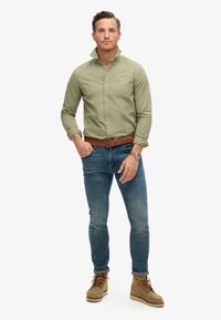 Camisa botonada verde claro con un bolsillo en el pecho, jeans azules y botas de cordones color beige. La camisa tiene el collar desabrochado y un diseño ajustado.