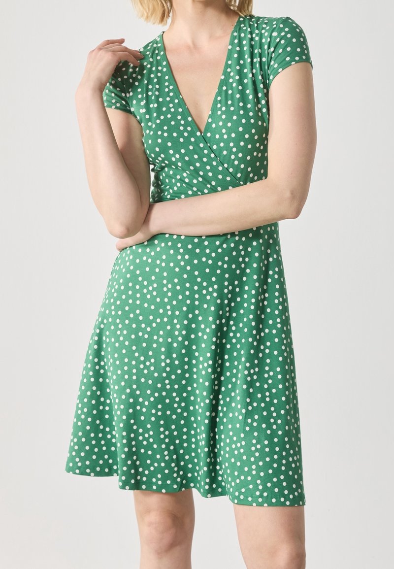 Femme portant une robe courte verte à pois blancs, manches courtes et col en V, debout avec un bras croisé et l'autre levé.