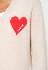 Beige kötött kardigán vörös szív grafikai mintával, amelyen a "FUTURE" felirat fehér színben szerepel. Az anyag puhanak és texturáltnak tűnik.