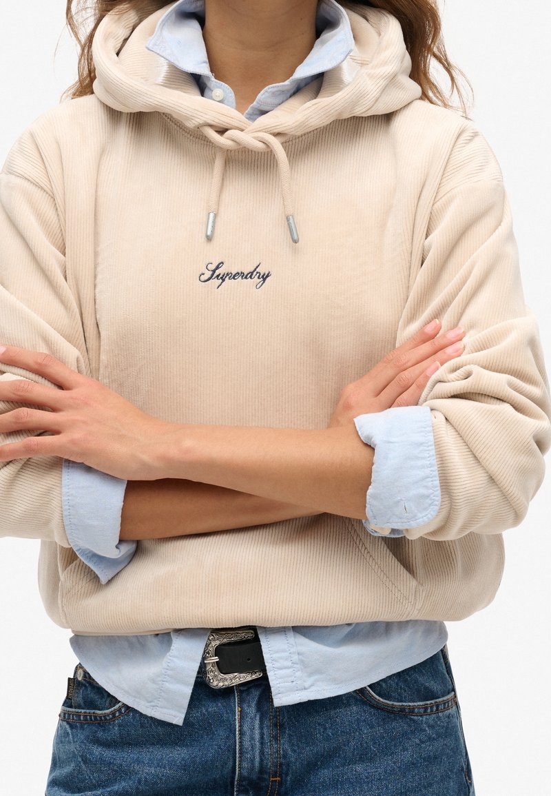 Felpa in corda beige con cappuccio regolabile, con il testo ricamato "Superdry" in blu navy. Indossata sopra una camicia azzurra a collare. Jeans di denim visibili.
