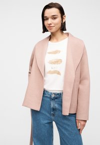 Νεαρή γυναίκα με κοντά σκούρα μαλλιά που φοράει ανοιχτό ροζ σακάκι, λευκό t-shirt με γραφικό φτερό και μπλε τζιν ψηλόμεσα, στέκεται μπροστά σε λευκό φόντο.